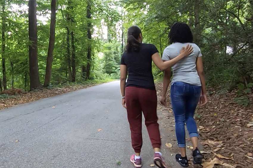 two teen girls walking together on a forest road, one putting her hand on the other's back in a gesture of support