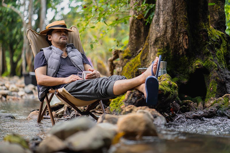A man dozes peacefully in a chair by a quiet river creek, surrounded by nature.