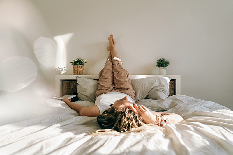 A woman relaxes on a bed, her legs comfortably propped up against the wall.