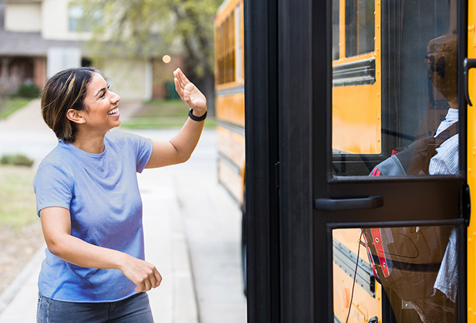 mother waving goodbye to child getting on a school bus